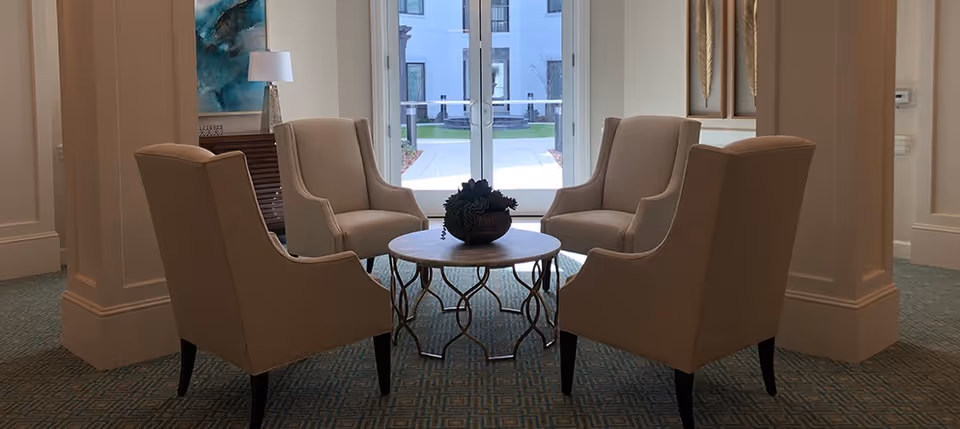 A seating area with four beige upholstered armchairs arranged around a round wooden table with a decorative plant centerpiece. The area is carpeted with a patterned design and is located near large glass doors leading outside. The walls are light-colored with decorative molding and artwork.