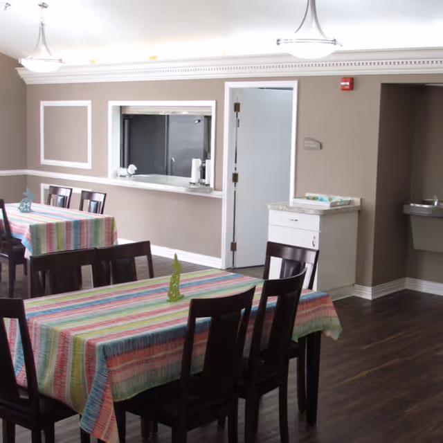 Dining room with tables covered in colorful striped tablecloths, dark wooden chairs, a serving window and a door set in beige walls.
