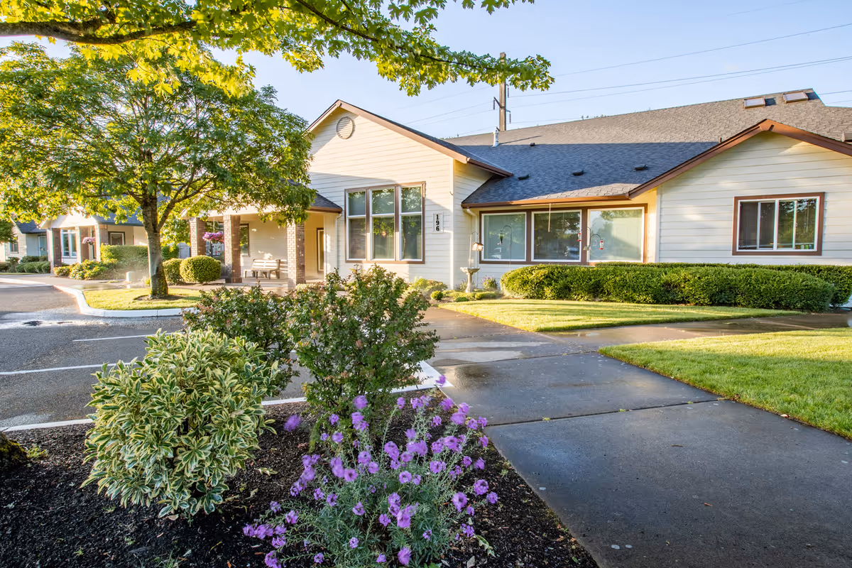 Exterior view of a single-story building with beige siding and brown trim, surrounded by well-maintained landscaping including bushes, purple flowers, and a tree. A paved walkway and parking area are visible in front of the building under a clear blue sky.