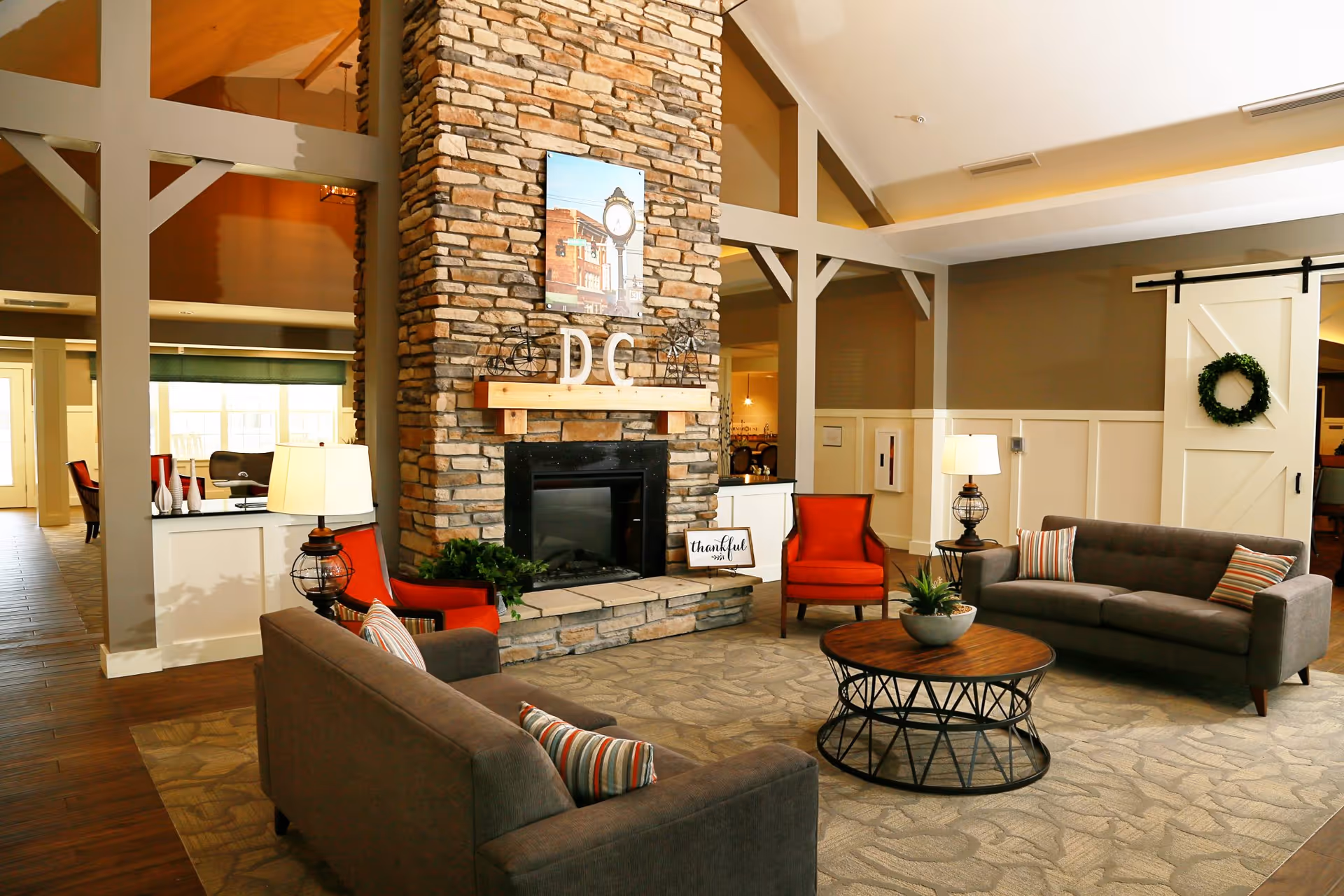 A cozy living room area with two gray sofas and two red armchairs arranged around a round wooden coffee table with a plant centerpiece. The room features a tall stone fireplace with a wooden mantle displaying decorative items including letters 'DC' and a small sign that says 'thankful'. The walls are painted in neutral tones with white wainscoting, and there is a white sliding barn door with a green wreath on it. The space is warmly lit with table lamps and natural light coming from windows in the background.