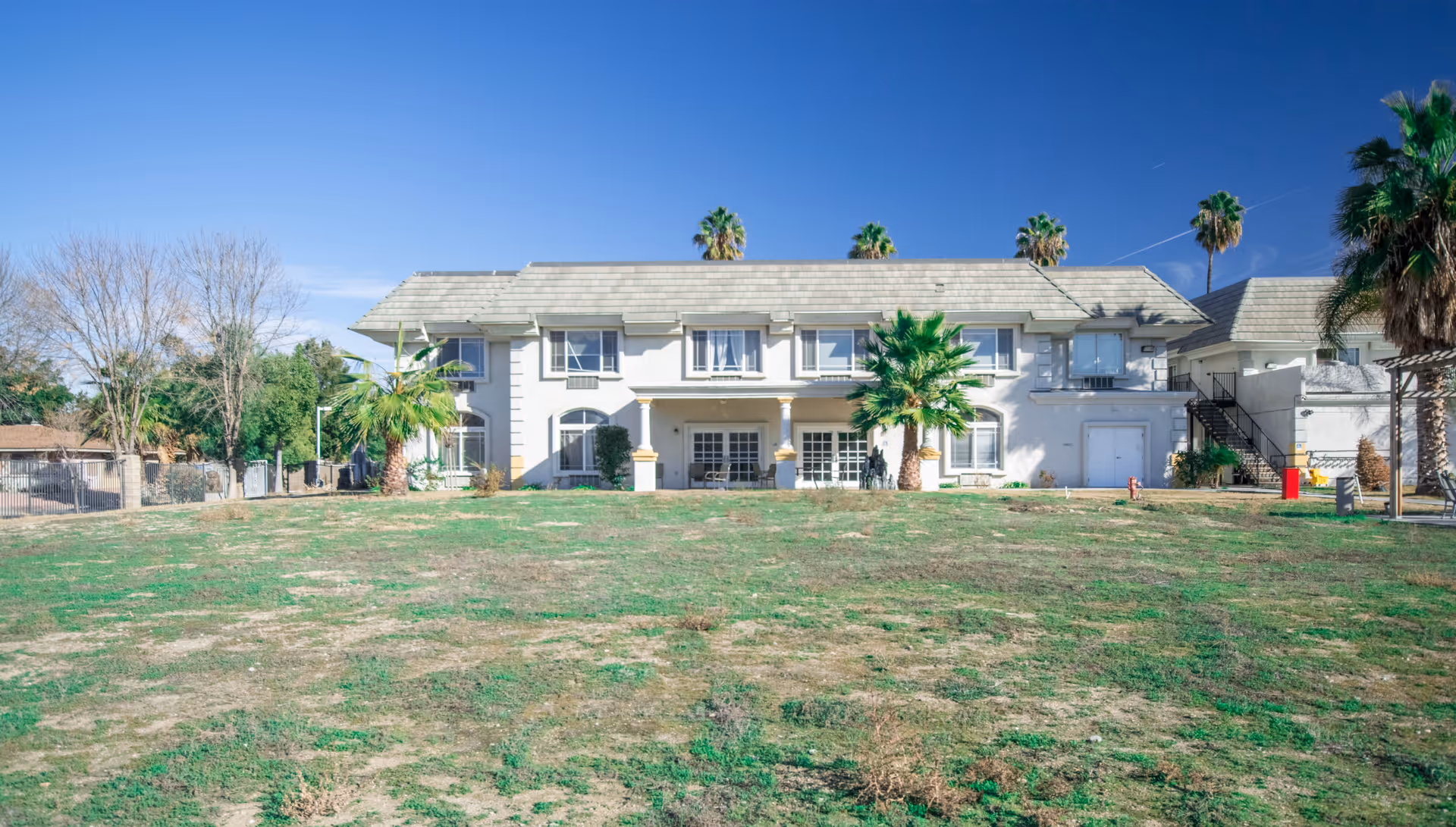 Exterior view of a two-story assisted living community building with a large grassy area in the foreground, palm trees around the building, and a clear blue sky above.