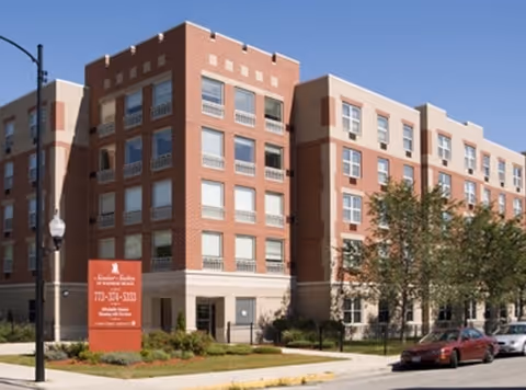 Exterior view of a multi-story senior living facility building with red brick and beige accents, surrounded by trees and parked cars on the street. A red sign in front displays the facility name and contact information.
