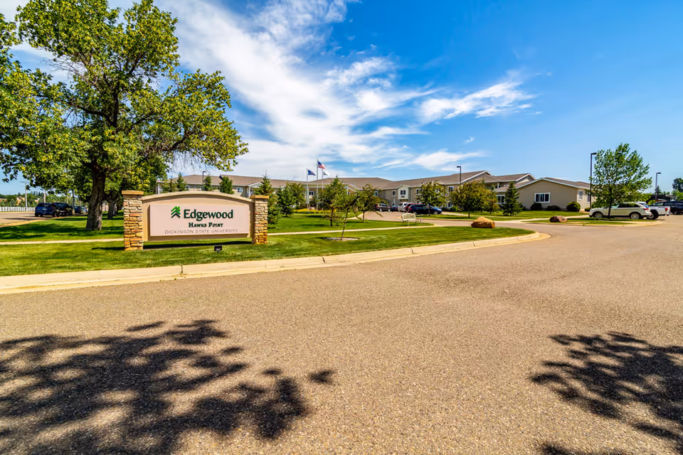 Exterior view of Edgewood Hawks Point facility with a large sign in the foreground, a paved driveway, green lawns, trees, parked cars, and a partly cloudy blue sky.