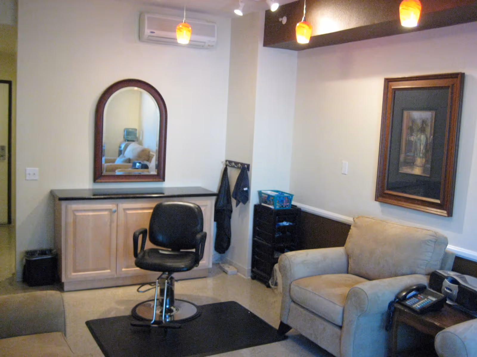 Interior room with a black salon chair on a black mat in front of a wooden cabinet with a mirror above it. There is a beige armchair to the right, a small table with a telephone and radio, and a framed picture on the wall. The room has light-colored walls and ceiling lights with orange pendant lamps.