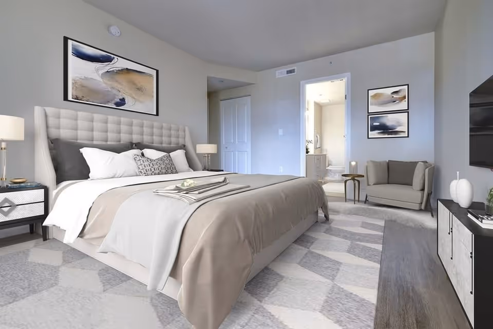 A modern bedroom with a large upholstered bed featuring beige and white bedding, two bedside tables with lamps, a small gray loveseat with a side table, abstract artwork on the walls, a patterned area rug, and a view into an adjoining bathroom.