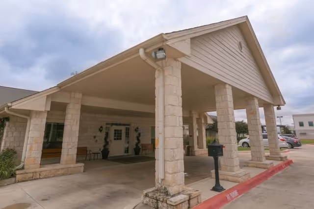 Entrance of Sonterra Health Center featuring a covered drop-off area supported by stone pillars, with a concrete driveway and parking spaces visible under a cloudy sky.