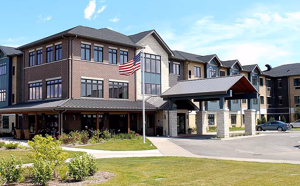 Exterior view of a multi-story senior living facility building with a covered entrance, an American flag on a flagpole, landscaped greenery, and a parking area with cars.