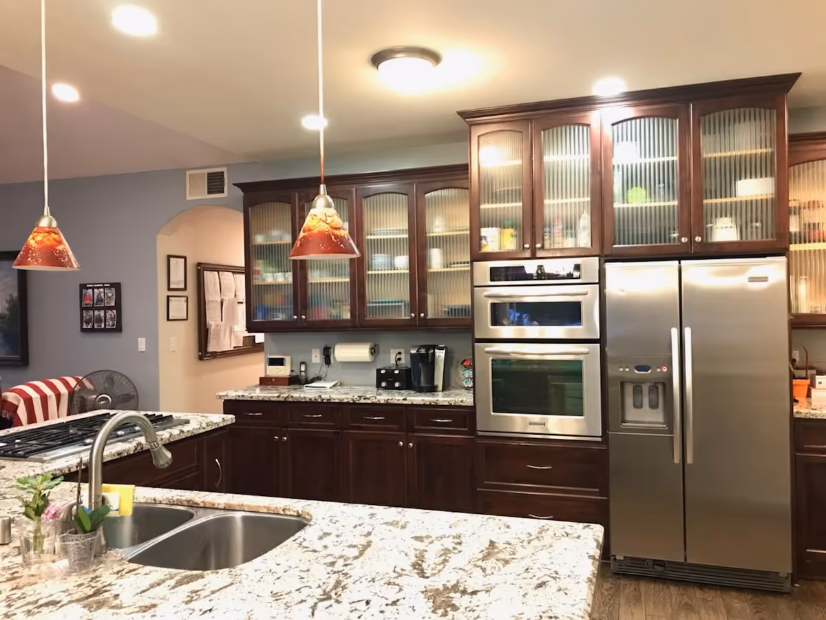A modern kitchen with dark wooden cabinets featuring frosted glass doors, a double stainless steel oven, and a large stainless steel refrigerator with water and ice dispenser. The kitchen has granite countertops with a double sink and a faucet. Two pendant lights with red and orange shades hang over the counter. In the background, there is a glimpse of a living area with a striped chair and a fan.