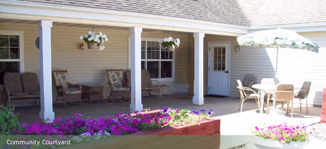 Covered outdoor patio area with seating including cushioned chairs and a table with an umbrella, surrounded by flower beds with purple and white flowers at Timberdale Trace Assisted Living.