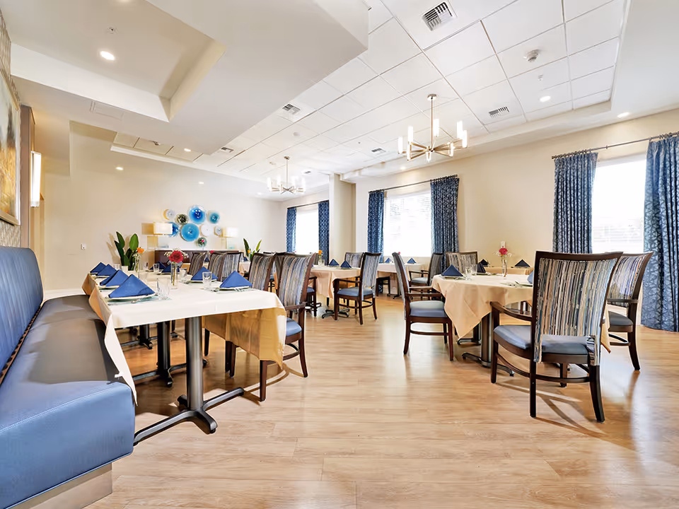 Bright, neatly arranged dining room with tables set with napkins and chairs, blue curtains, and decorative wall art.