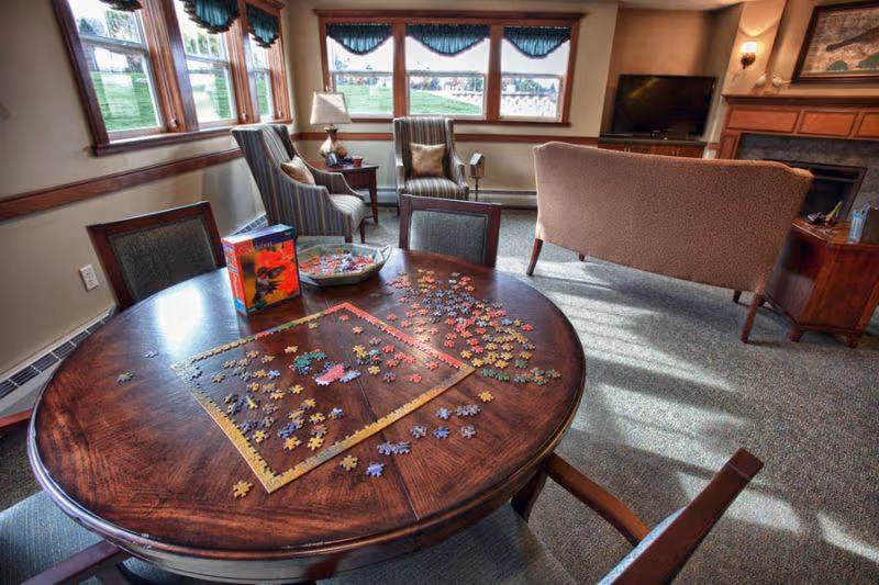 Sunlit common living area with a round table strewn with jigsaw puzzle pieces, chairs, a sofa, TV, and windows.
