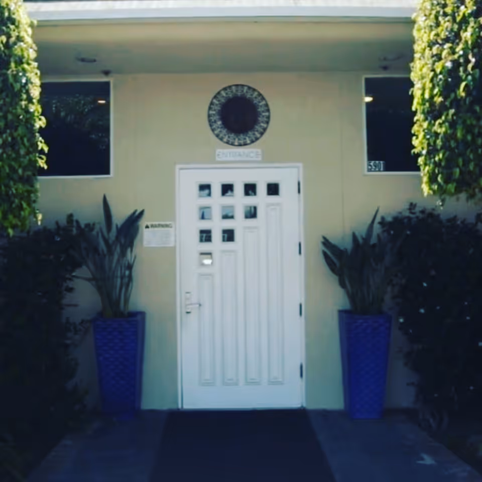 Front entrance of a building with a white door featuring small square windows at the top. The door is flanked by two tall blue planters with green plants. There are two windows on either side of the door, and a circular decorative element above the door with the word 'ENTRANCE' below it. Green foliage partially frames the entrance.