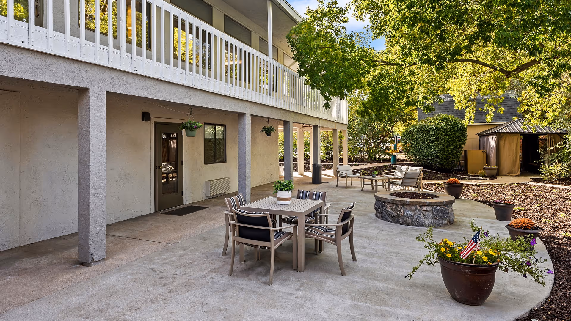 Outdoor patio area at a retirement community with a table and four chairs, a stone fire pit surrounded by additional seating, potted plants including one with small American flags, and trees providing shade. The building has a white railing balcony above and beige walls with a door and window below.