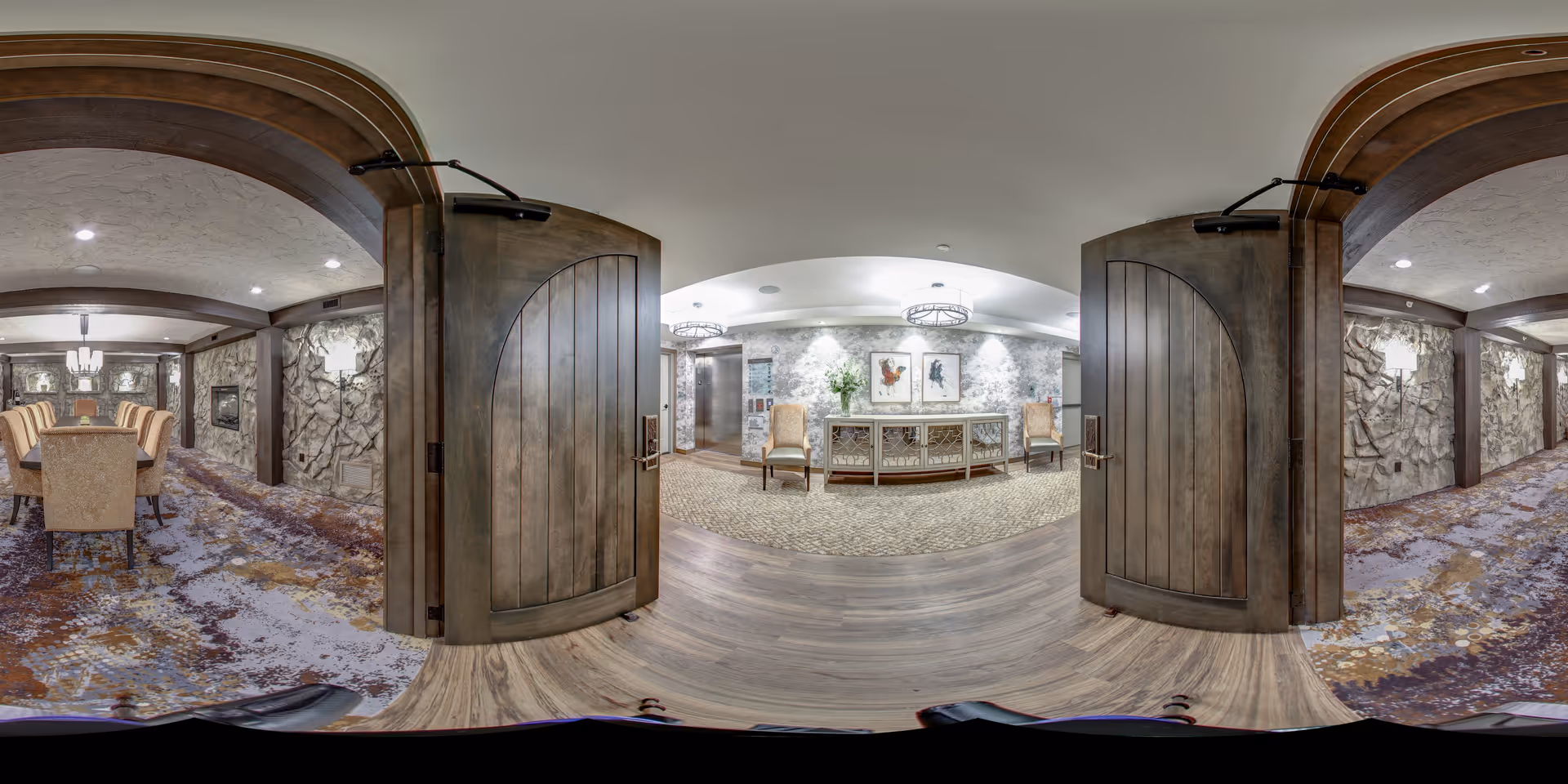 Open wooden double doors lead into a decorated senior living common area with seating, a dining table to the left, a console table with artwork, and textured stone walls.