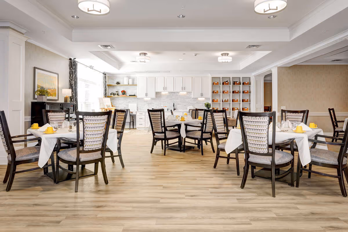 A bright and spacious dining room with several round and rectangular tables covered with white tablecloths. Each table is set with yellow cups and glassware. The room features wooden chairs with patterned cushions, light wood flooring, white cabinetry, and shelves with decorative items. Large windows with curtains allow natural light to fill the space.