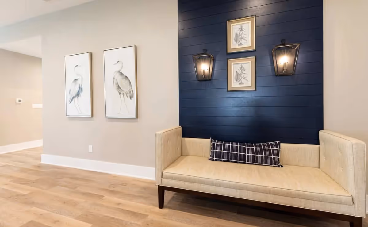 A cozy seating area with a beige upholstered bench featuring a black and white checkered pillow. Behind the bench is a dark blue shiplap accent wall with two wall-mounted lantern-style lights and three framed botanical prints. To the left, two framed bird illustrations hang on a light beige wall. The floor is light wood.