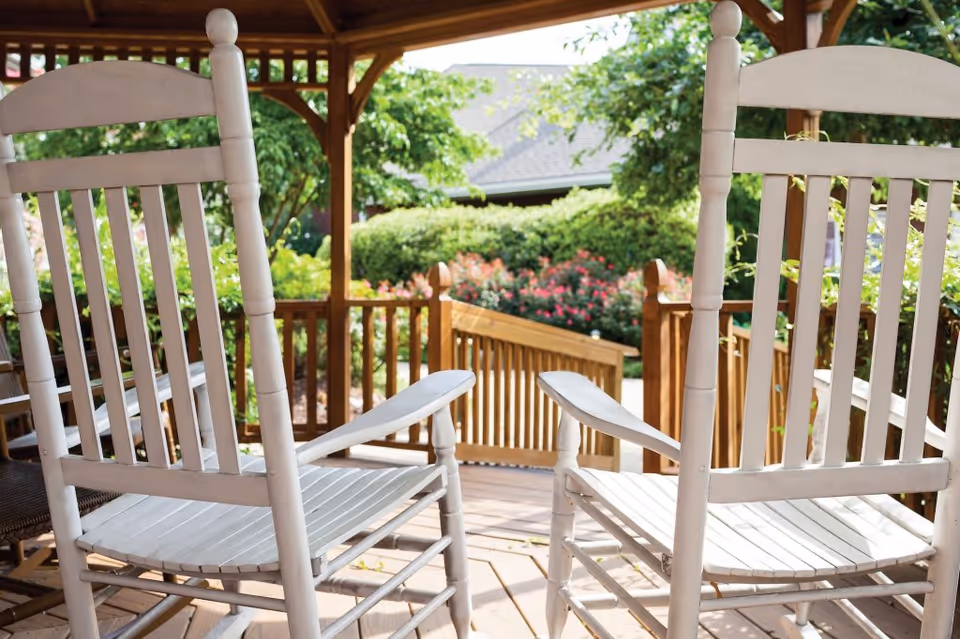 Two white wooden rocking chairs on a wooden porch with a railing, surrounded by greenery and flowering bushes under a wooden gazebo roof.
