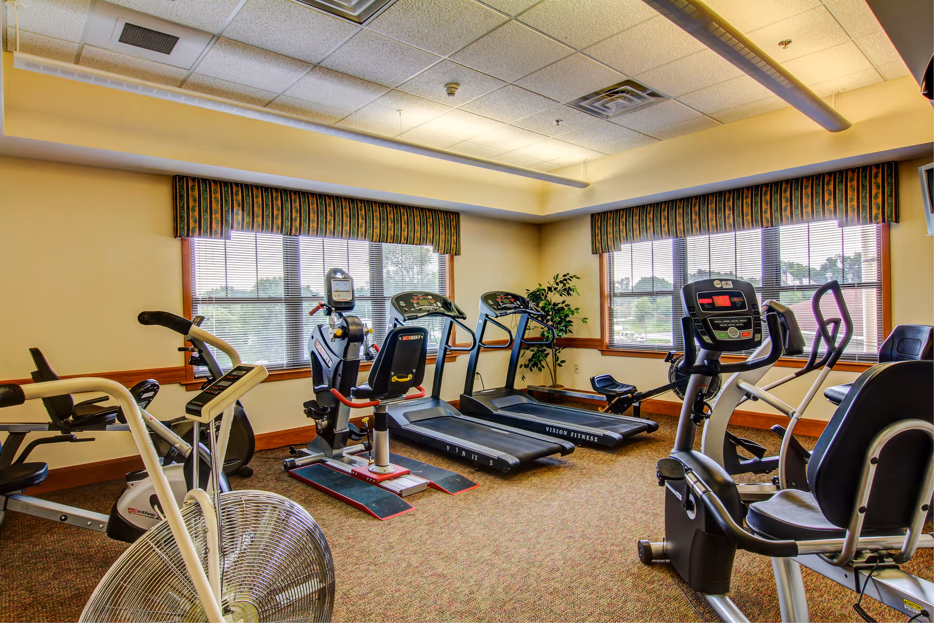 A fitness room with various exercise equipment including stationary bikes, treadmills, and an elliptical machine. The room has large windows with blinds and patterned valances, beige walls, and a carpeted floor. There is a potted plant in the corner and fluorescent lighting on the ceiling.