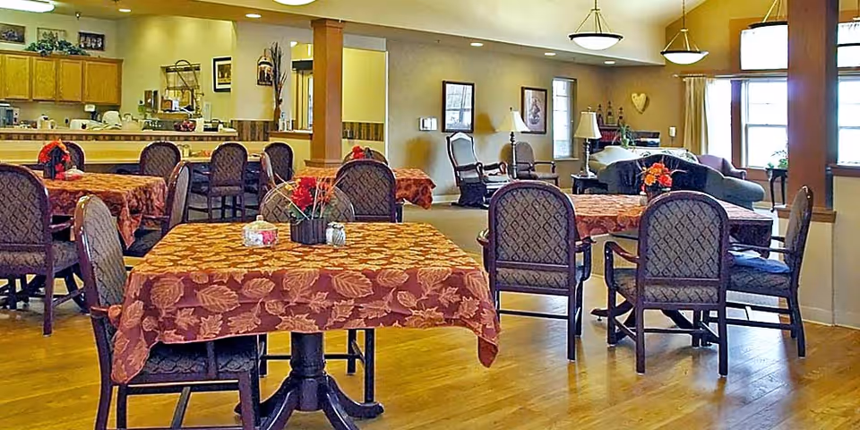 A warm and inviting dining area in a senior living facility with several tables covered in autumn leaf-patterned tablecloths. Each table has a small floral centerpiece. The room features wooden floors, cushioned chairs, and soft lighting from ceiling fixtures. In the background, there is a kitchen counter area and a cozy sitting area with armchairs and a sofa near windows with curtains.
