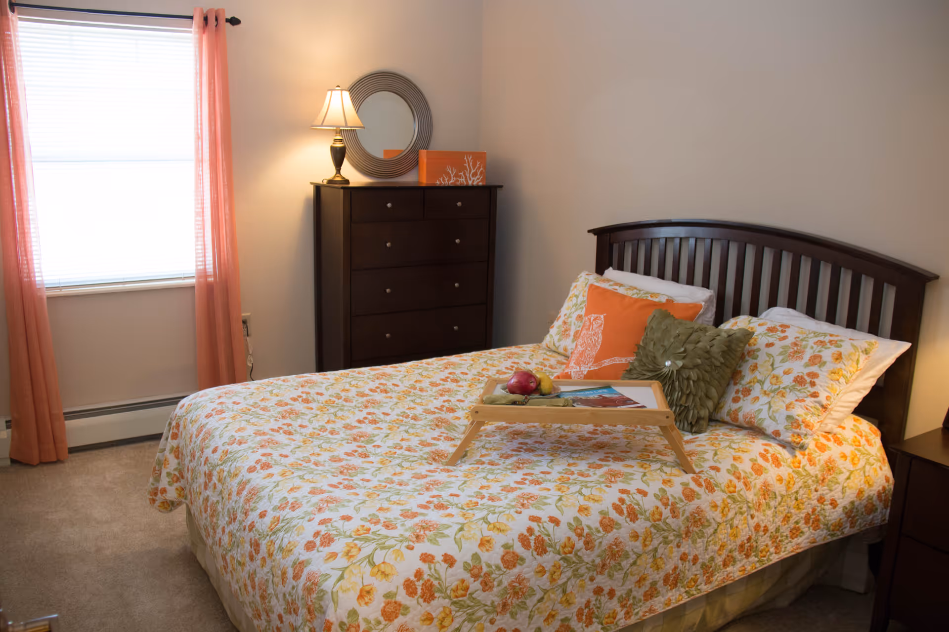 A cozy bedroom with a bed covered in a floral-patterned quilt with orange, yellow, and green flowers. The bed has three pillows, including an orange pillow with an owl design and a green textured pillow. A wooden tray with fruit and a magazine sits on the bed. To the left of the bed is a window with pink curtains, and next to the window is a dark wooden dresser with a round mirror and a table lamp on top. The walls are light-colored, and the floor is carpeted.