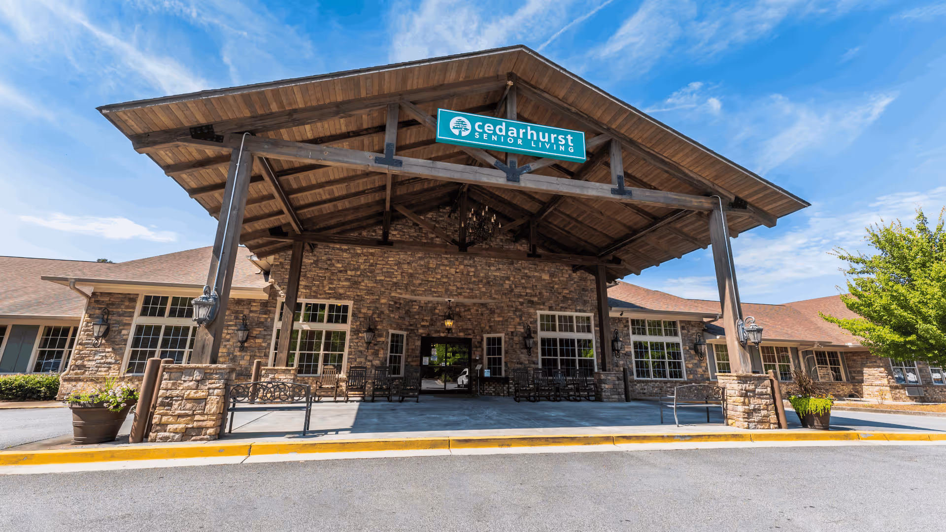 Front exterior view of Cedarhurst Senior Living of Canton showing a large covered entrance with stone walls, wooden beams, benches, and a sign with the facility name under a blue sky.