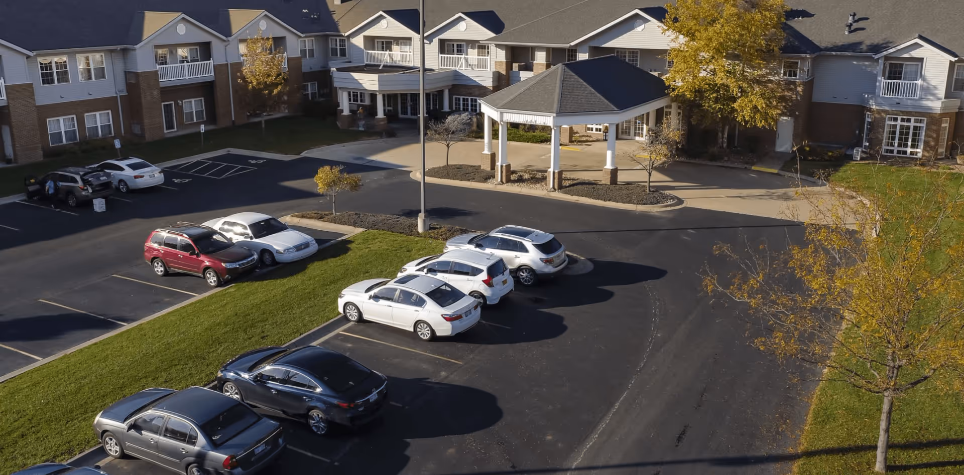 Aerial view of the front entrance and parking lot of a senior living facility with several parked cars, a covered drop-off area, and a multi-story building with balconies and windows surrounded by trees and grass.