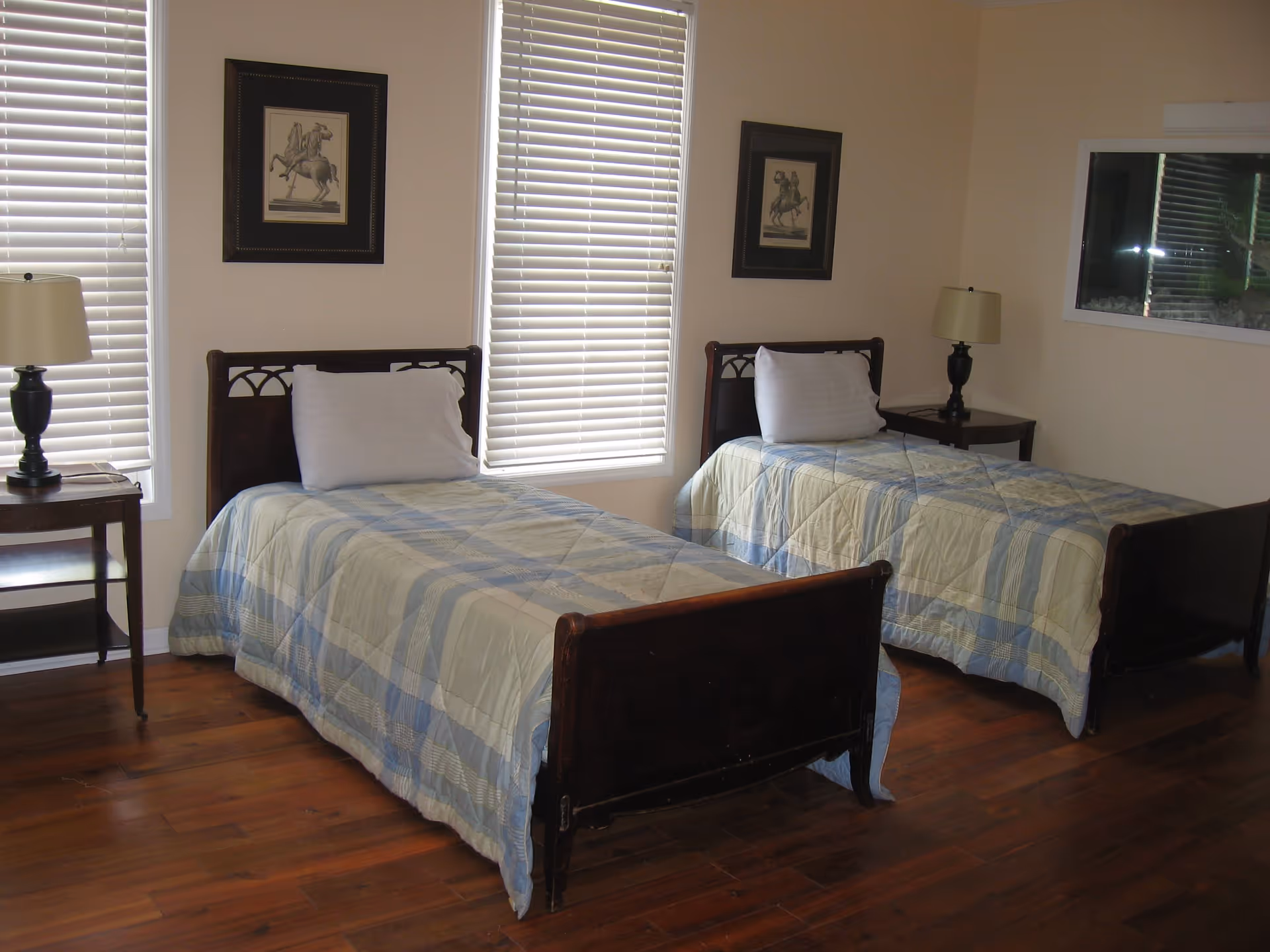 A bedroom with two single beds placed parallel to each other, each with a blue and beige plaid bedspread and a white pillow. There are two windows with white blinds behind the beds, two framed pictures of horse riders on the wall, and two bedside tables with lamps. The floor is wooden.