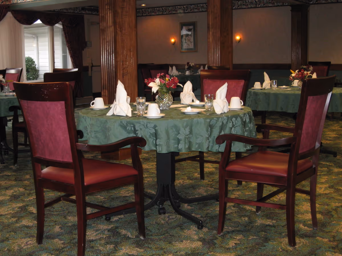A dining room with round tables covered in green tablecloths, each set with white folded napkins, cups, glasses, and small flower arrangements. There are wooden chairs with red cushioned seats and backs around the tables. The room has carpeted floors and wooden columns, with framed artwork and wall sconces on the walls.