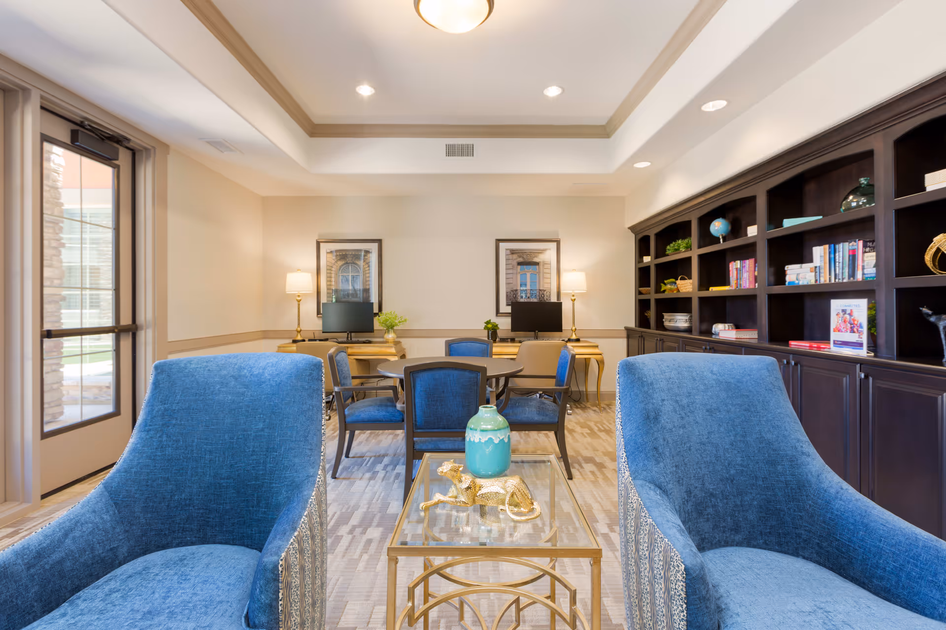 A cozy senior living common area with two blue upholstered armchairs in the foreground, a glass-top coffee table with decorative items, a round table with four blue chairs in the middle, and a dark wood built-in bookshelf filled with books and decor on the right. Two desks with computers and lamps are against the back wall, which is decorated with framed artwork. A glass door on the left lets in natural light.