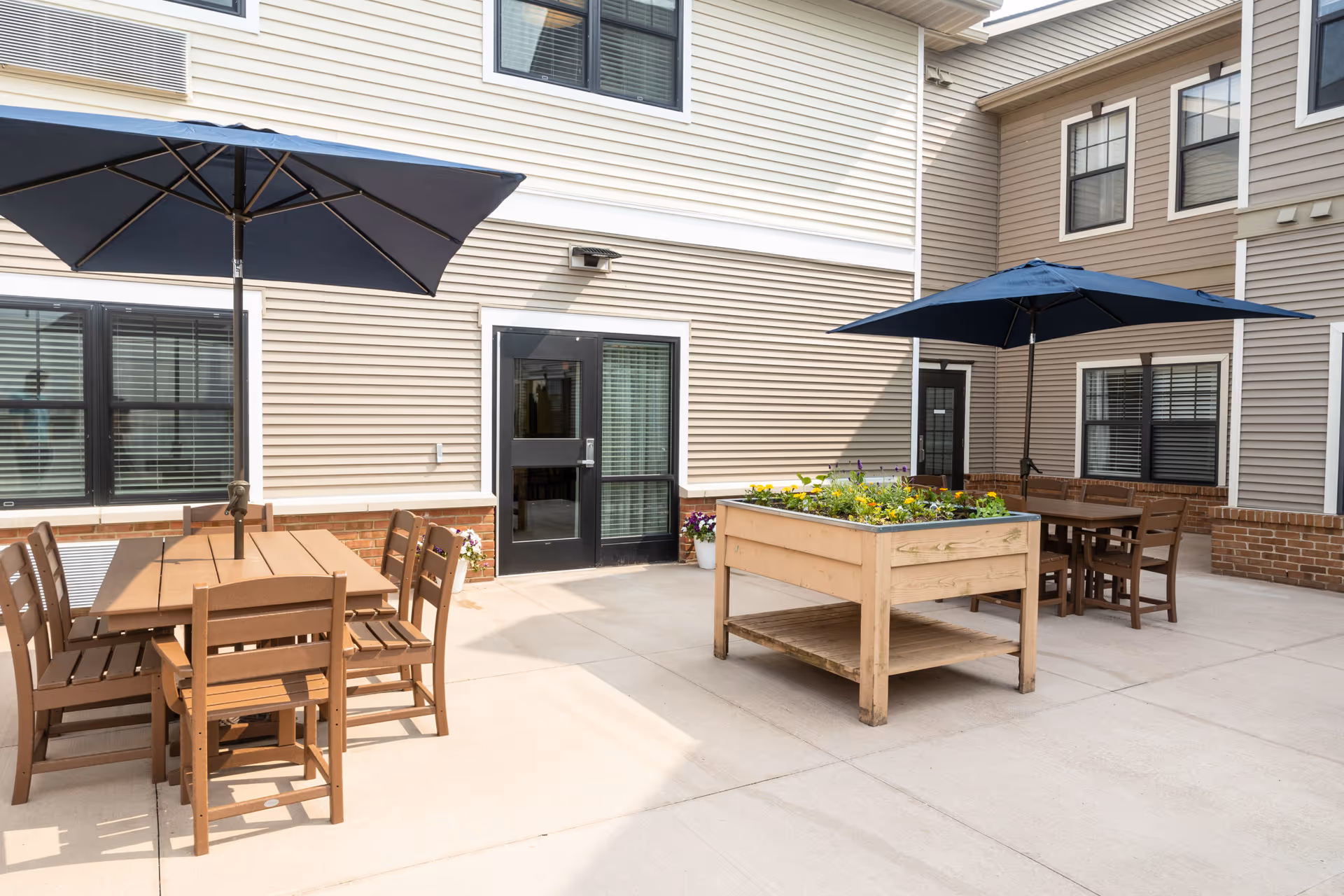 Outdoor courtyard patio with wooden tables and chairs, blue umbrellas, and a raised planter in front of a multi-story building.