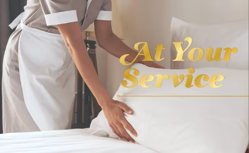 A person wearing a gray uniform and white apron is making a bed with white sheets and pillows. The text 'At Your Service' is overlaid in gold lettering.