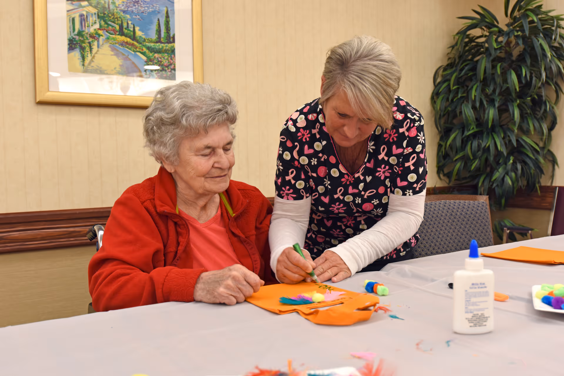 An elderly woman in a red jacket sitting at a table while a caregiver in a patterned scrub top helps her with a craft project involving orange paper, glue, and colorful pom-poms in a room with beige walls, a framed painting, and a large green plant.