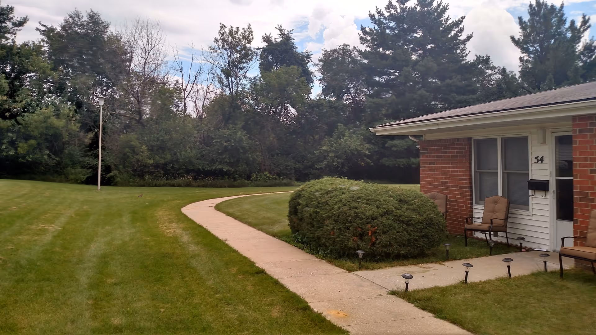 A curved concrete walkway leading to the entrance of a single-story brick and white siding building with the number 54. The entrance has two cushioned chairs on either side and a mailbox. The building is surrounded by a well-maintained grassy lawn, bushes, and tall trees in the background under a partly cloudy sky.
