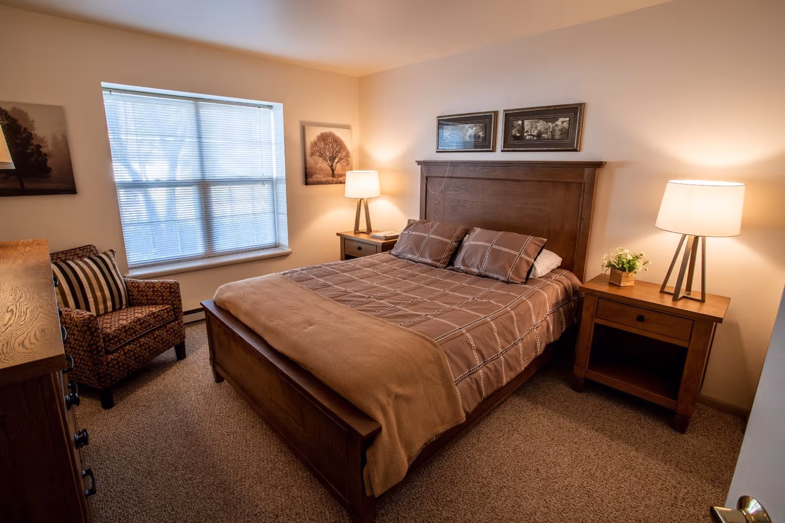 A cozy bedroom with a wooden bed frame and a neatly made bed with brown checkered bedding. There are two matching wooden nightstands on either side of the bed, each with a lamp and one with a small plant. A patterned armchair with a striped pillow is placed near a window with blinds. The walls are decorated with framed pictures of trees.