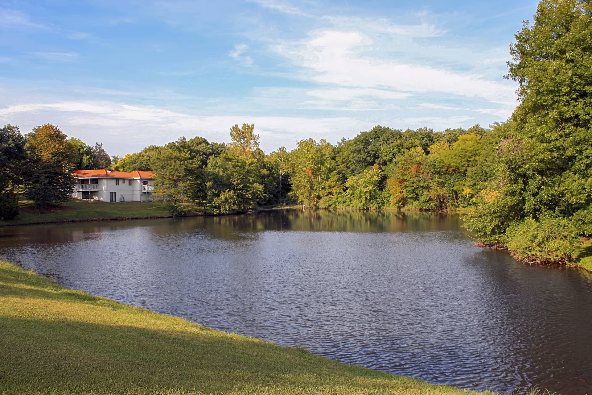 A peaceful outdoor scene featuring a calm pond surrounded by green trees and grass, with a building with a red roof partially visible on the left side under a partly cloudy sky.