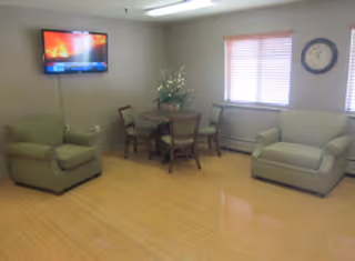 A small common area with light wood flooring, two green upholstered armchairs, a round wooden table with four chairs, a wall-mounted flat screen TV, a large clock on the wall, and two windows with blinds partially closed.