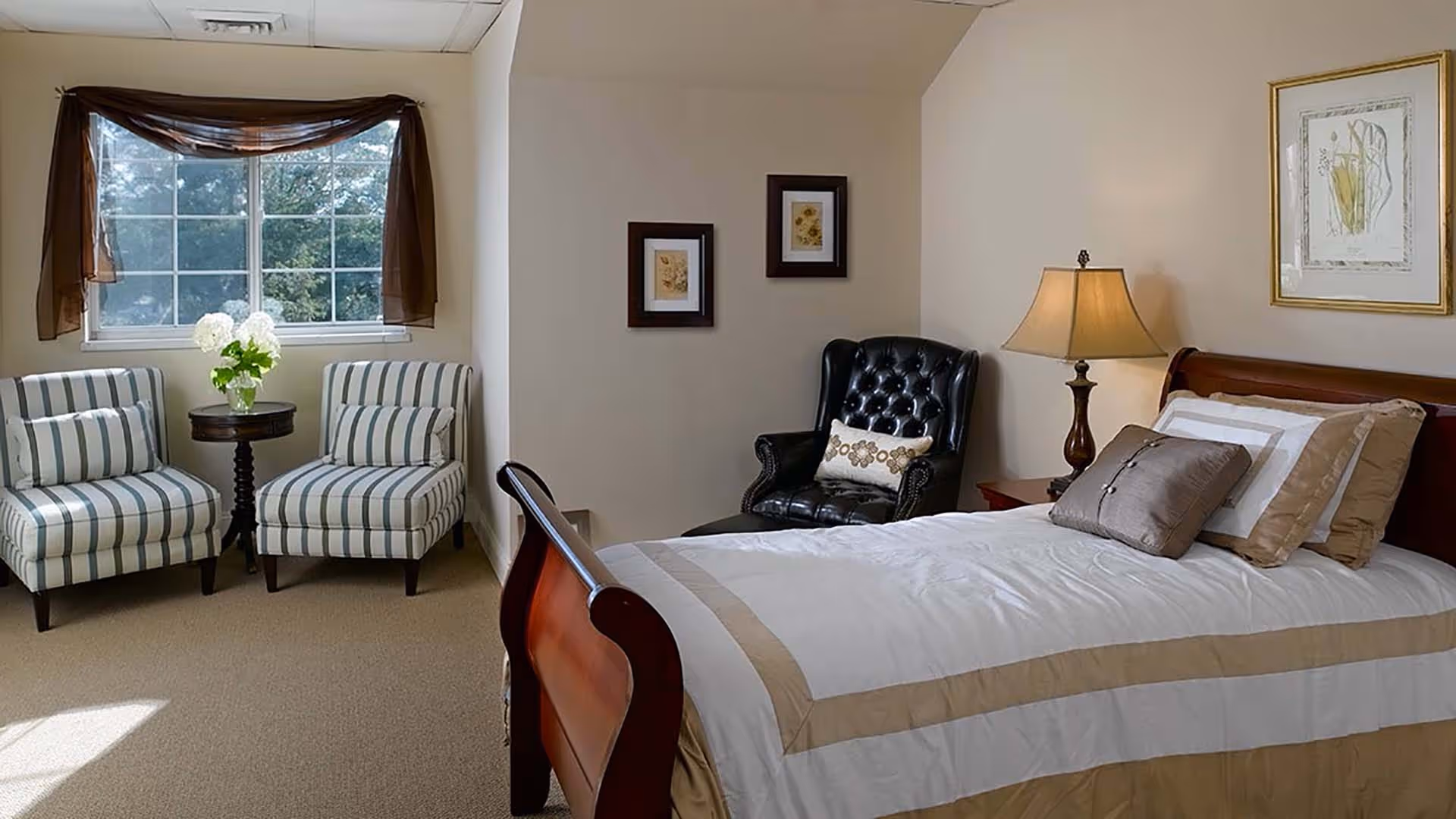 A cozy bedroom with a wooden sleigh bed made with white and beige bedding, a bedside table with a lamp, a black leather armchair, two striped armchairs with a small round table between them holding a vase of white flowers, and a window with brown sheer curtains.