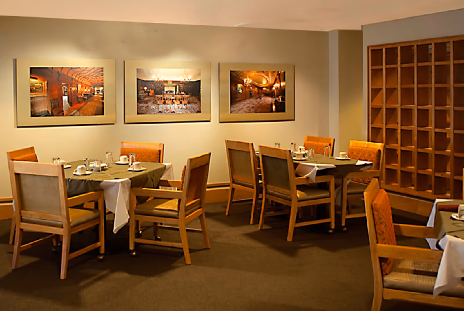 A dining room with several wooden tables and chairs arranged neatly. Each table is set with cups, saucers, and condiments. The walls are decorated with three framed pictures, and there is a wooden shelving unit on the right side of the room.