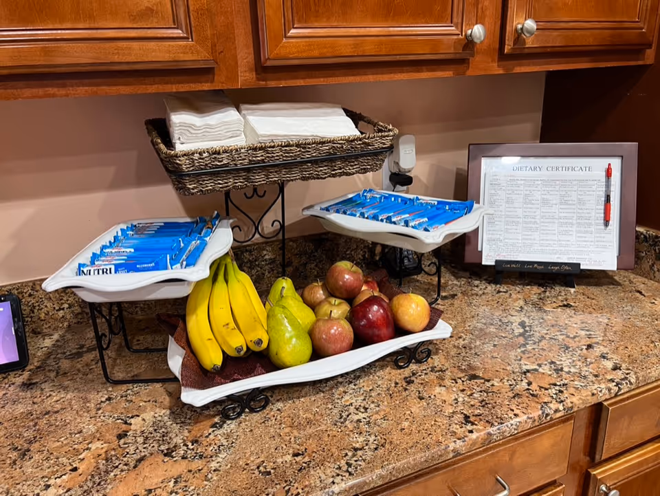 A kitchen counter with a three-tiered serving tray holding bananas, pears, apples, and Nutri-Grain bars. Above the counter are wooden cabinets, and to the right is a framed dietary certificate with a pen attached. A basket with napkins is on the top tier of the serving tray.