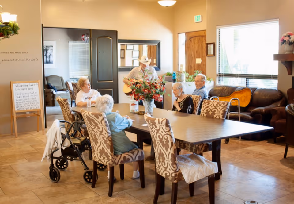 A group of elderly people sitting around a large table in a well-lit common room at Heritage Manor Assisted Living. One person is playing a guitar while others listen. The room has patterned chairs, a walker, a couch, and a large window letting in natural light. There are flowers on the table and a whiteboard with notes in the background.