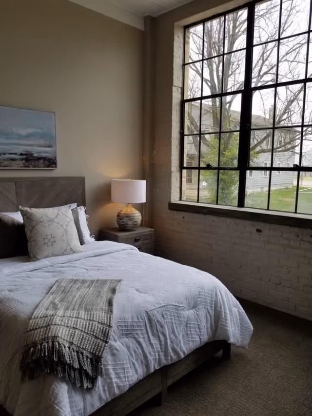 A cozy bedroom with a neatly made bed featuring white bedding and a decorative pillow. A textured throw blanket is draped over the foot of the bed. Next to the bed is a wooden nightstand with a ceramic table lamp. A large window with black metal framing lets in natural light and offers a view of trees and a neighboring building outside. A painting hangs on the wall above the headboard.
