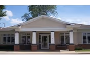Front exterior view of a single-story building with beige siding, white trim, and brick accents. The entrance has a covered porch supported by columns, and there are multiple windows on either side of the door. The sky is partly cloudy.