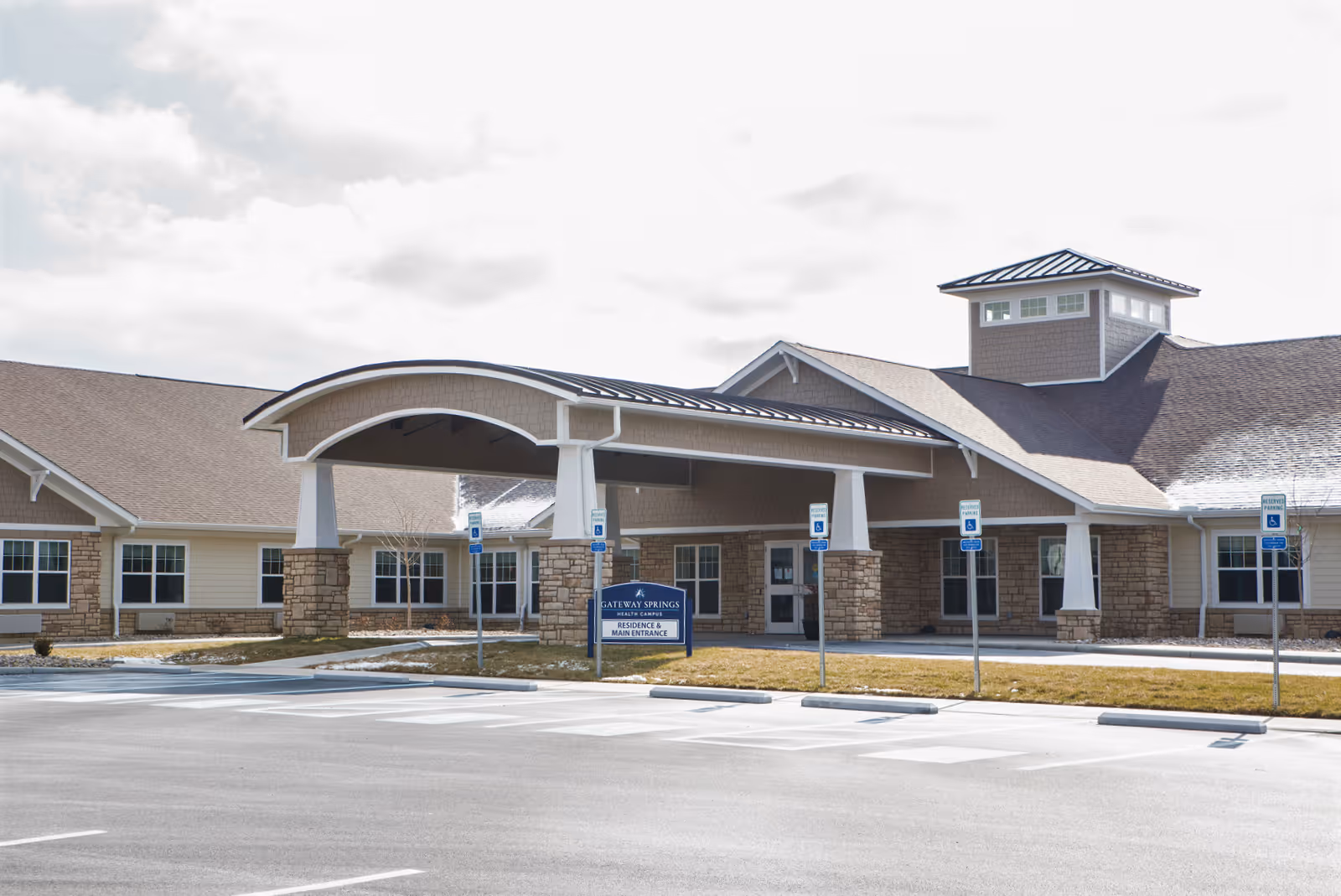 Exterior view of Gateway Springs Health Campus showing the main entrance with a covered drop-off area supported by stone pillars. The building has a combination of stone and siding with multiple windows and a tower-like structure on the roof. Several handicap parking spaces are visible in front of the entrance.
