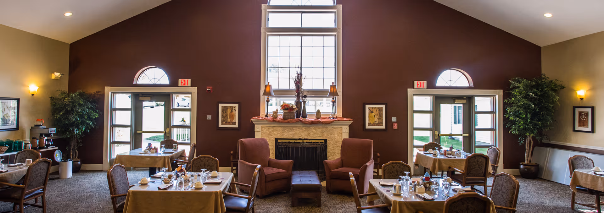 A cozy dining room with several tables covered in beige tablecloths, set with cups, glasses, and silverware. The room features a fireplace in the center with two armchairs and a small ottoman in front of it. Large windows and glass doors allow natural light to enter, and there are plants and framed artwork on the walls.