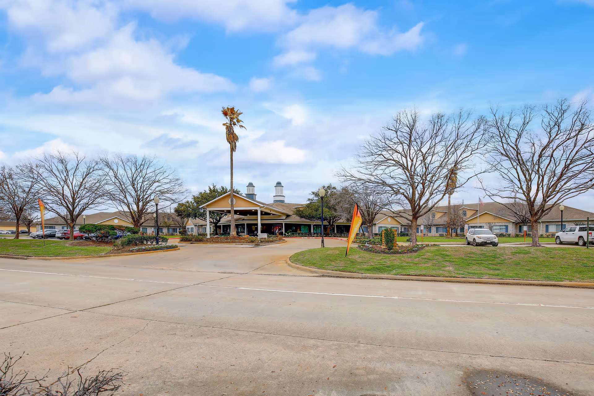 Exterior view of Morada Cy-Fair senior living facility showing a large driveway entrance, landscaped grassy areas with leafless trees, a tall palm tree, and several parked cars under a partly cloudy sky.