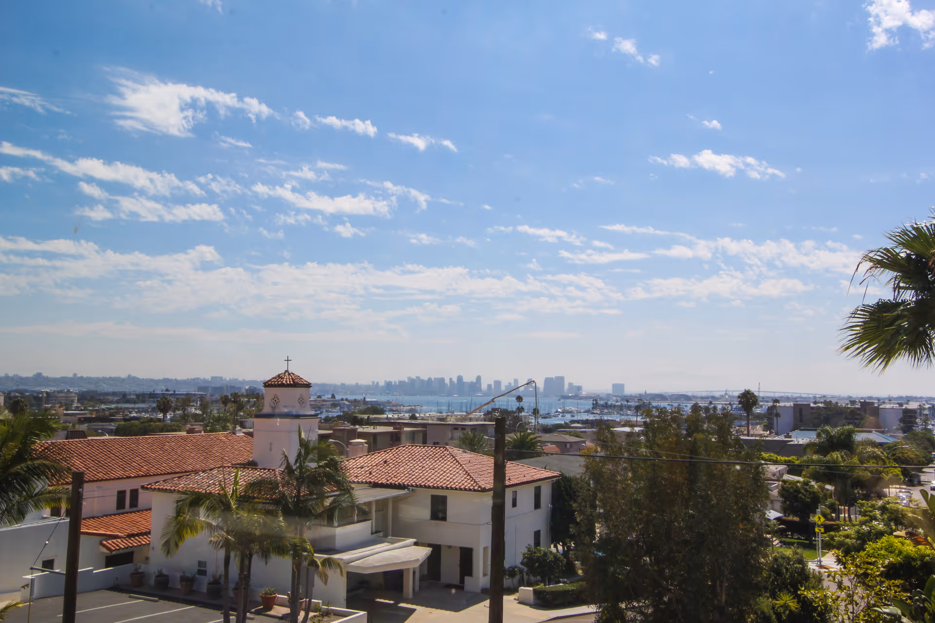 View of a residential area with a building featuring a red-tiled roof and a small tower with a cross on top, palm trees, and a distant city skyline under a partly cloudy blue sky.