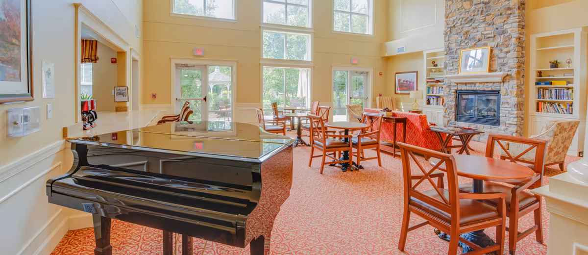 Bright senior community lounge with a black grand piano, multiple tables and chairs, a stone fireplace with built-in bookshelves, and large windows.