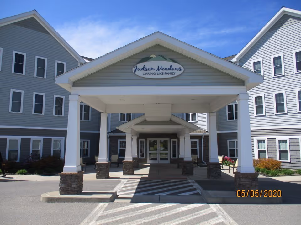 Front entrance of Judson Meadows Assisted Living facility with a covered drop-off area supported by white columns with stone bases. The building is light gray with white trim and multiple windows. The sky is clear and blue.