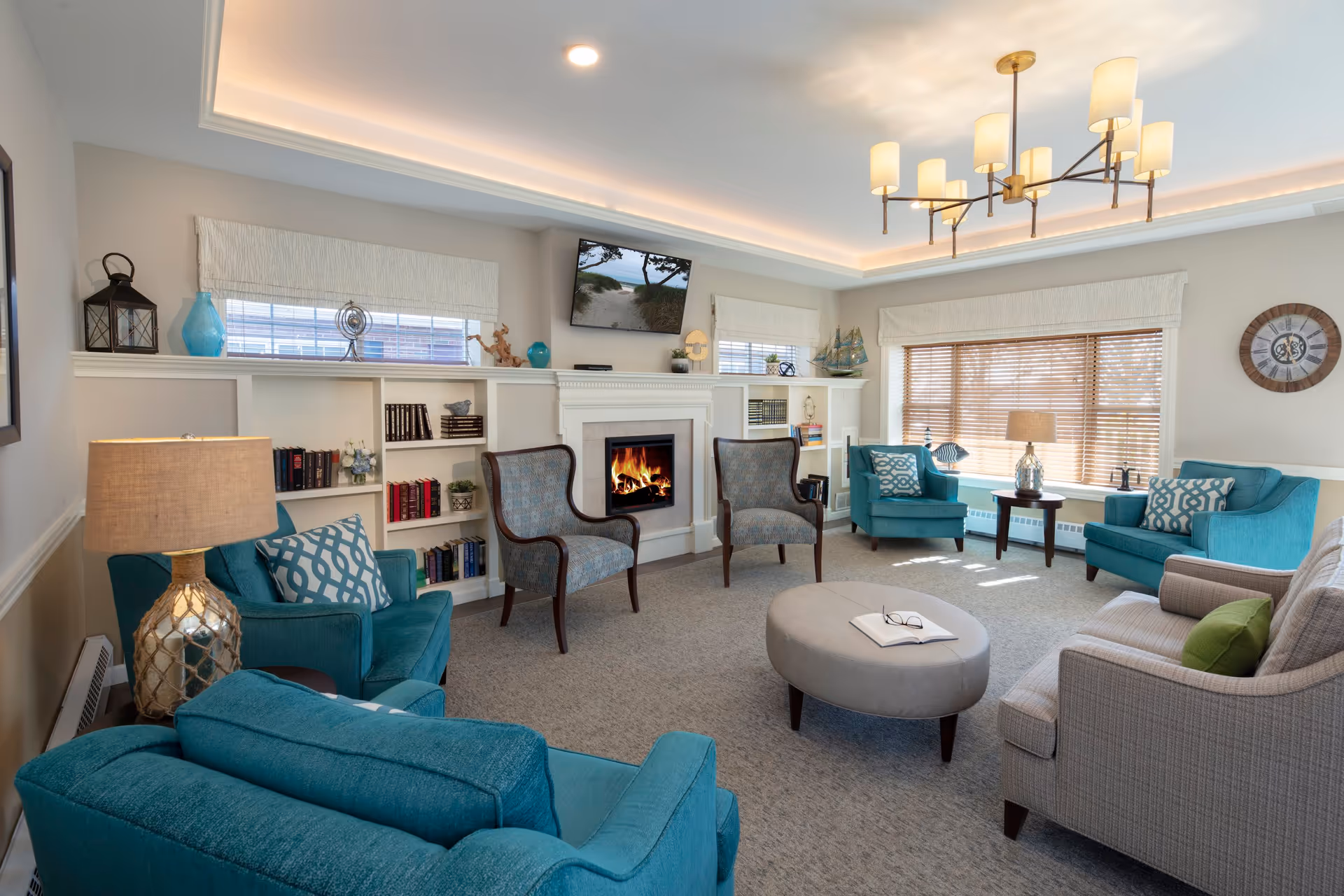 A cozy living room with teal and beige armchairs arranged around a gray oval ottoman with an open book and glasses on it. The room features a fireplace with a fire burning, built-in white shelves with books and decorative items, a wall-mounted TV above the fireplace, large windows with blinds, a round wall clock, and a modern chandelier with multiple lamps.