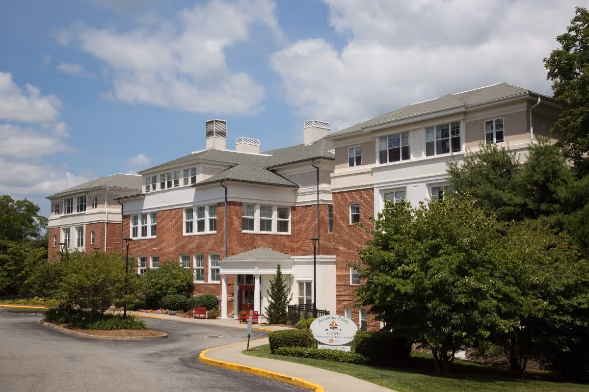 Exterior view of a multi-story senior living facility building with red brick and white accents, surrounded by trees and landscaping under a partly cloudy sky. A sign near the entrance reads 'Academy Point at Mystic.'